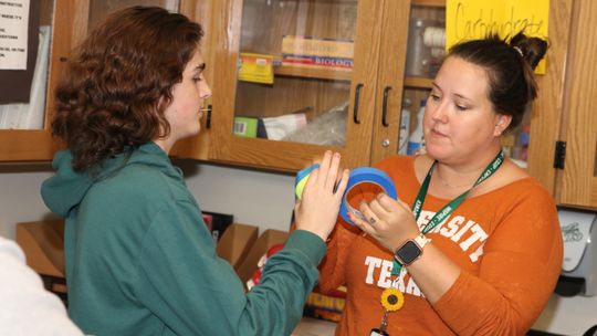 Legacy biology teacher Julie Hubbard tapes a tennis ball to a student’s hand to represent a competitive inhibitor during a lab on enzymes.