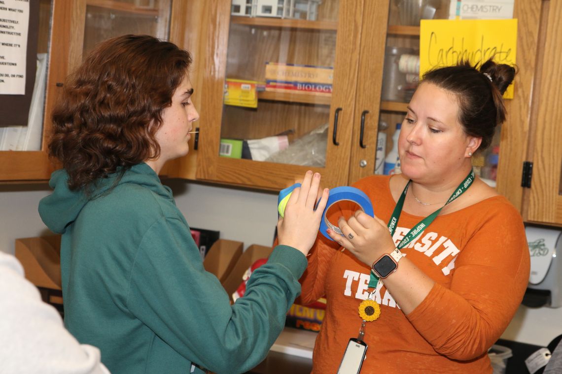 Legacy biology teacher Julie Hubbard tapes a tennis ball to a student’s hand to represent a competitive inhibitor during a lab on enzymes.